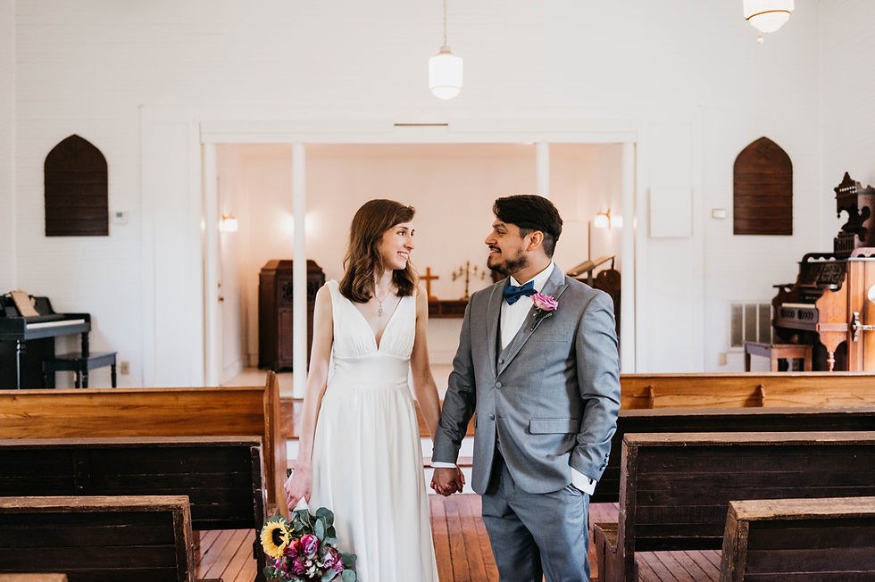A bride and groom holding hands in the aisle of the Chapel, between wooden pews, with the pump organ and piano behind them.