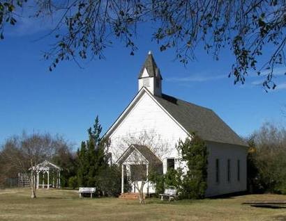 The Chapel: a white clapboard church with a small steeple, framed by tree branches, with benches and a gazebo on the lawn.