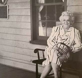Lera Millard Thomas, smiling, seated on a bench on the porch of a clapboard house.