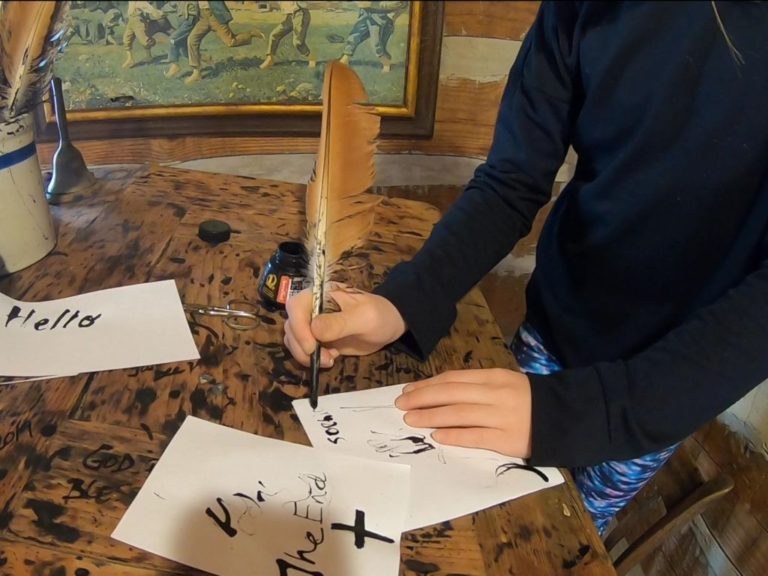 A student practices writing with a feather quill and a bottle of ink at a worn wooden desk.